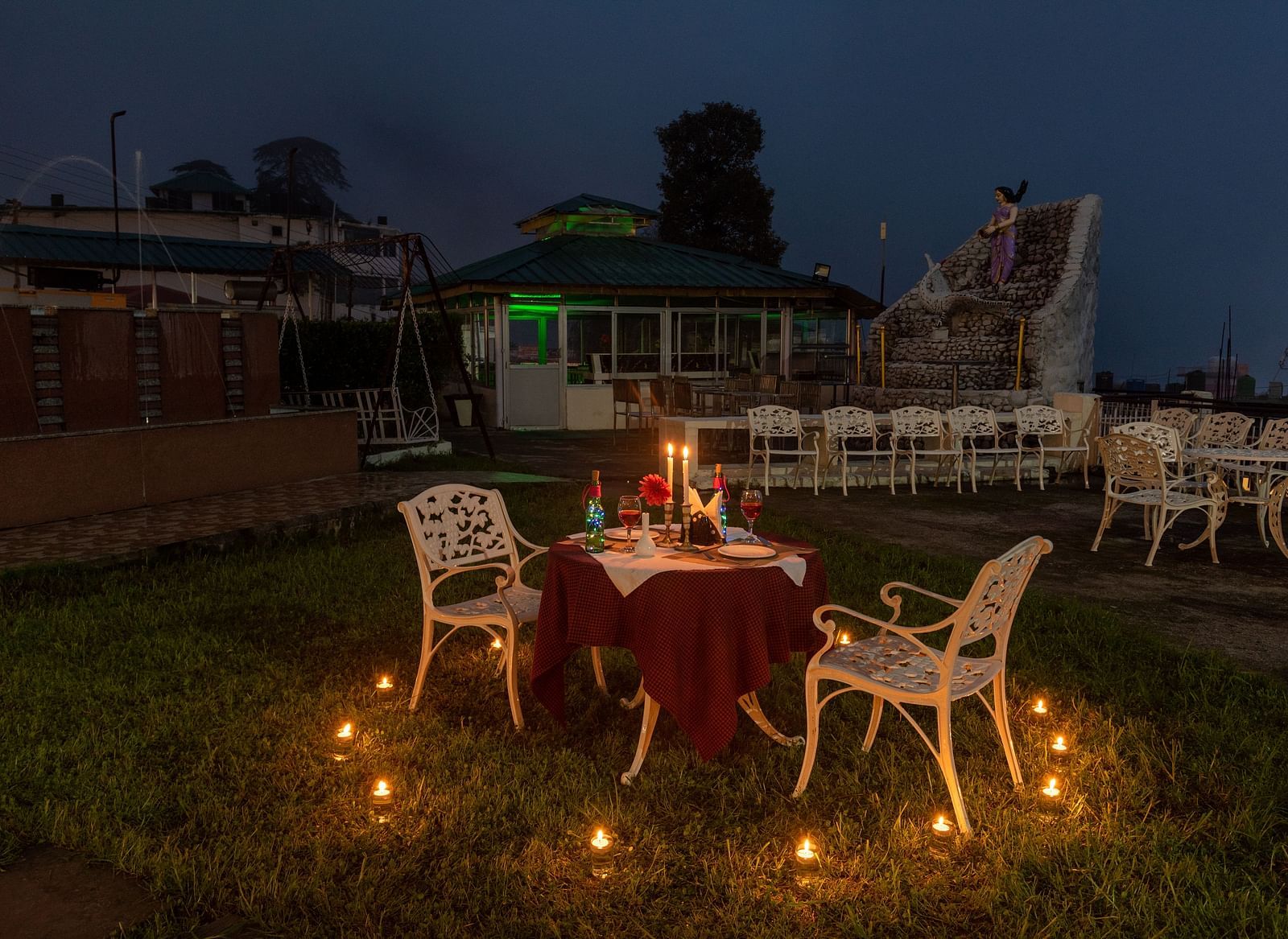 Candlelight dinner setup with a table for two, surrounded by candles and a view of a small fountain and seating area in the background - DLS Meghavan Resort, Dharamshala
